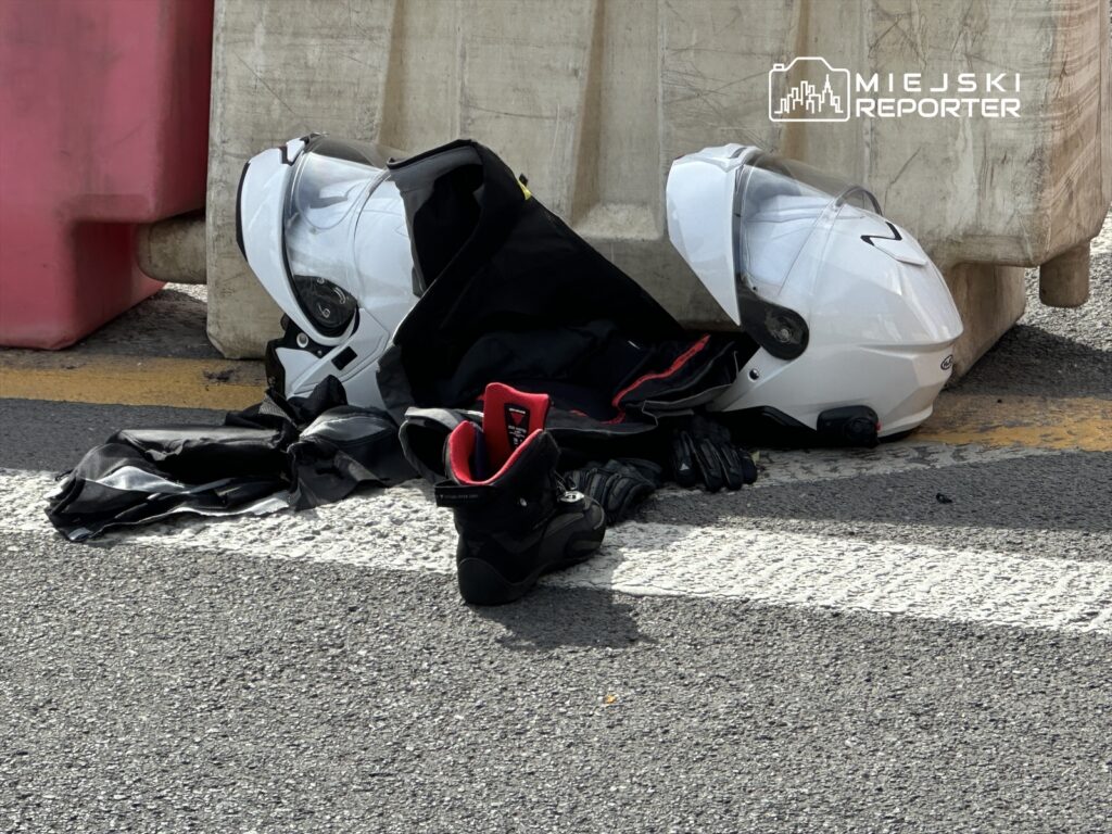 Na drodze leżą dwa białe kaski, czarna kurtka motocyklowa oraz rękawice i buty. W tle widoczna bariera ochronna.