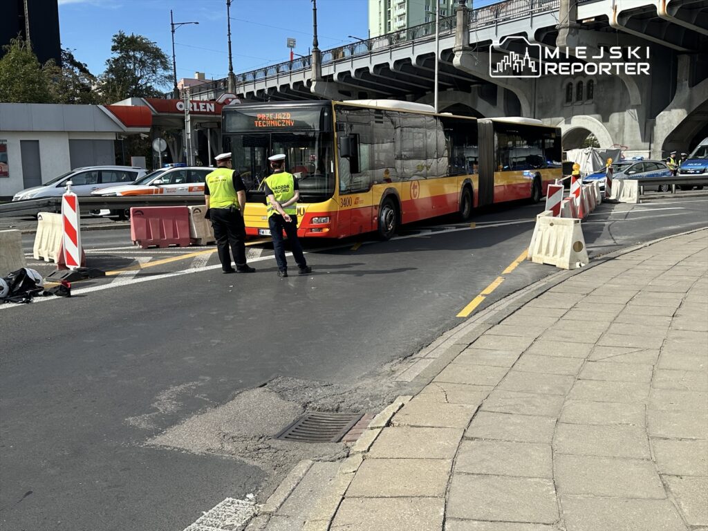 Dwaj policjanci w odblaskowych kamizelkach rozmawiają obok autobusu z napisem "Przejazd techniczny" na ruchliwej ulicy.