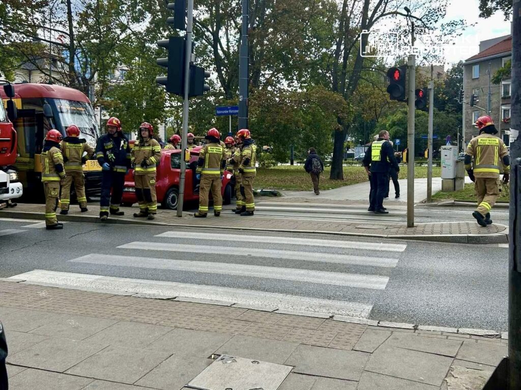 Grupa strażaków w hełmach i mundurach omawia sytuację obok czerwonego samochodu na ulicy, w tle widać tramwaj.