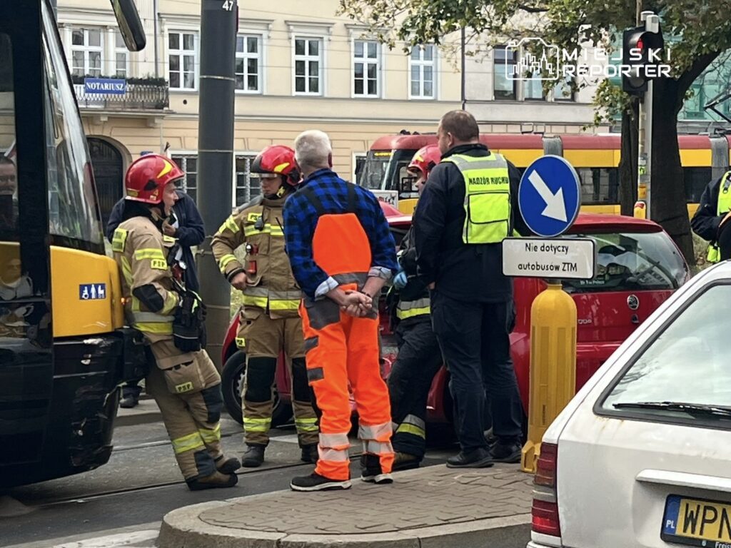 Grupa strażaków w mundurach rozmawia z mężczyzną w pomarańczowej kamizelce na ruchliwej ulicy.