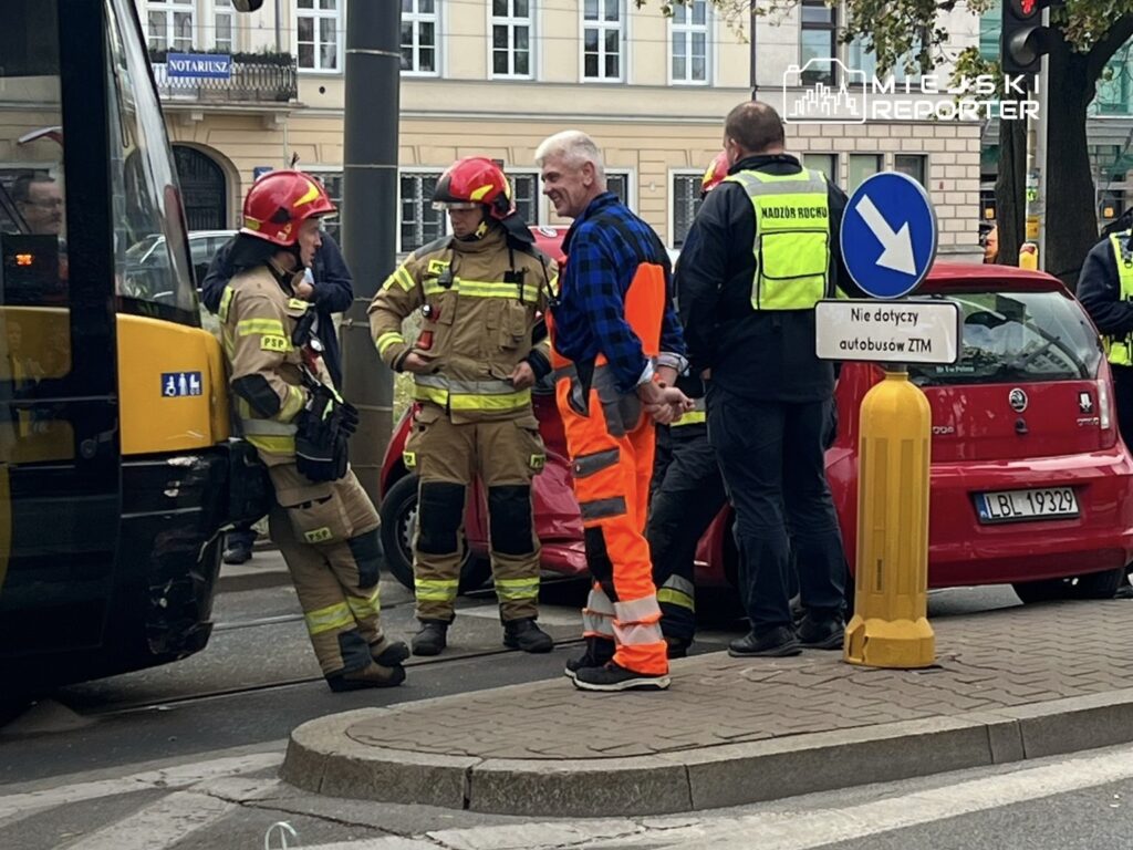 Grupa strażaków w mundurach rozmawia z mężczyzną w pomarańczowych spodniach na ulicy obok autobusu.