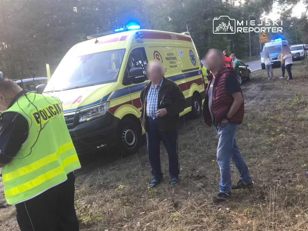 Policjant w kamizelce odblaskowej rozmawia z dwoma mężczyznami obok karetki pogotowia w lesie.