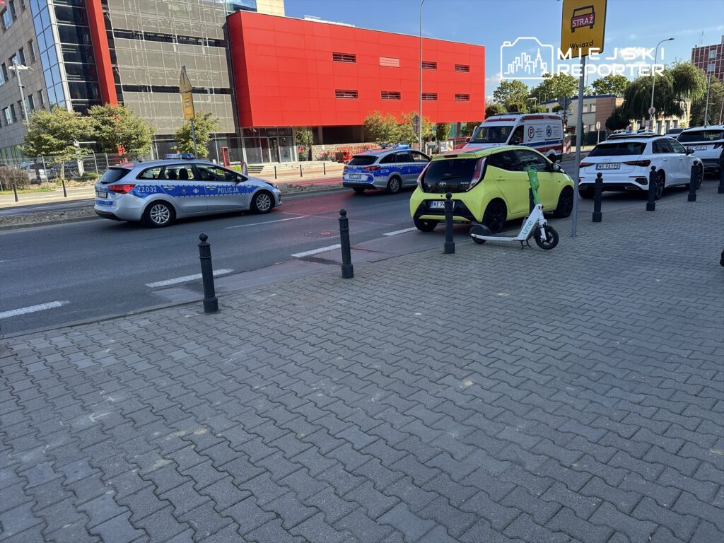 Policjant w radiowozie przejeżdża obok nowoczesnych budynków, a na chodniku stoi elektryczna hulajnoga.