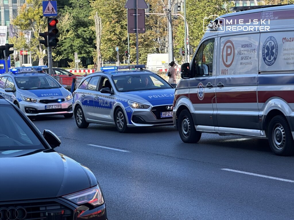 Policyjny radiowóz z niebieskimi światłami zatrzymany na skrzyżowaniu, obok karetka pogotowia. W tle widać zielone drzewa.