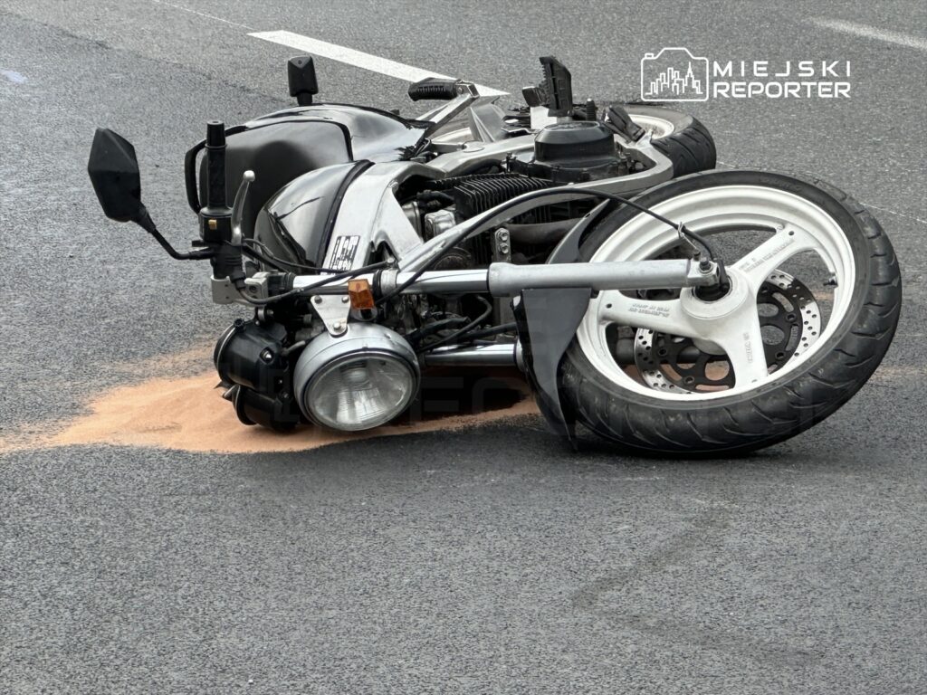 Motocykl leżący na boku na drodze, z plamą piasku obok, w miejskim otoczeniu.