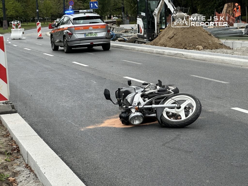 Motocykl leży na boku na jezdni, obok oznakowania budowy oraz radiowóz z włączonymi sygnałami świetlnymi.