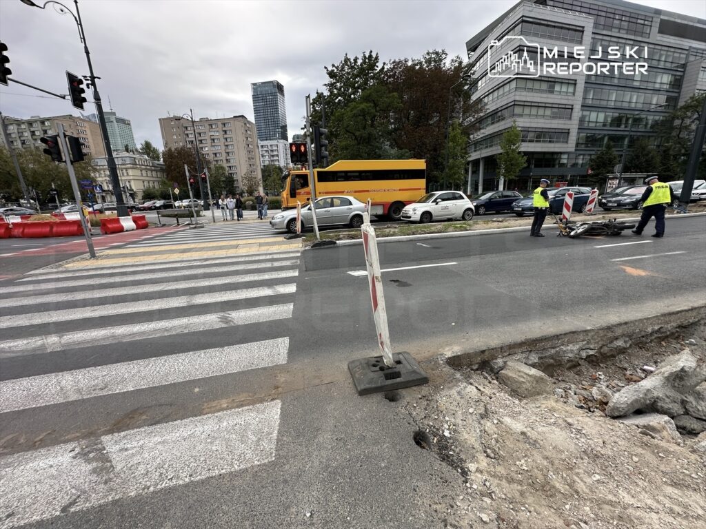 Grupa osób na przejściu dla pieszych obserwuje prace drogowe, podczas gdy policjanci zabezpieczają miejsce zdarzenia.