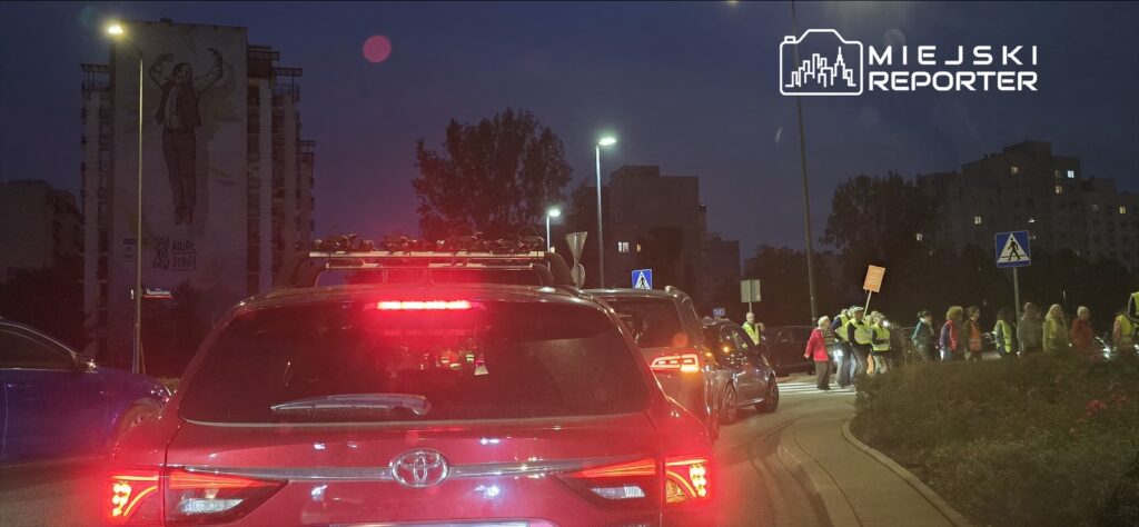 Grupa osób w odblaskowych kamizelkach idzie wzdłuż drogi, a samochody stoją w korku w tle.