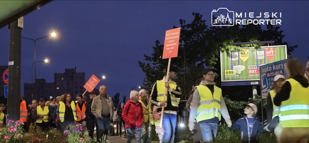Grupa osób w kamizelkach odblaskowych idzie chodnikiem, trzymając transparenty, w tle widoczne billboardy.