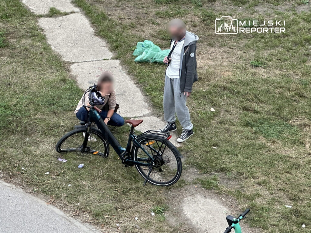 Kobieta w jasnej bluzie naprawia rower na trawniku, obok stoi mężczyzna w szarej bluzie i dresach.