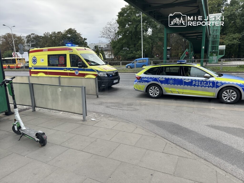 Ambulans z włączonymi sygnałami świetlnymi zatrzymany obok radiowozu policji na miejskiej ulicy. Na chodniku stoi elektryczna hulajnoga.