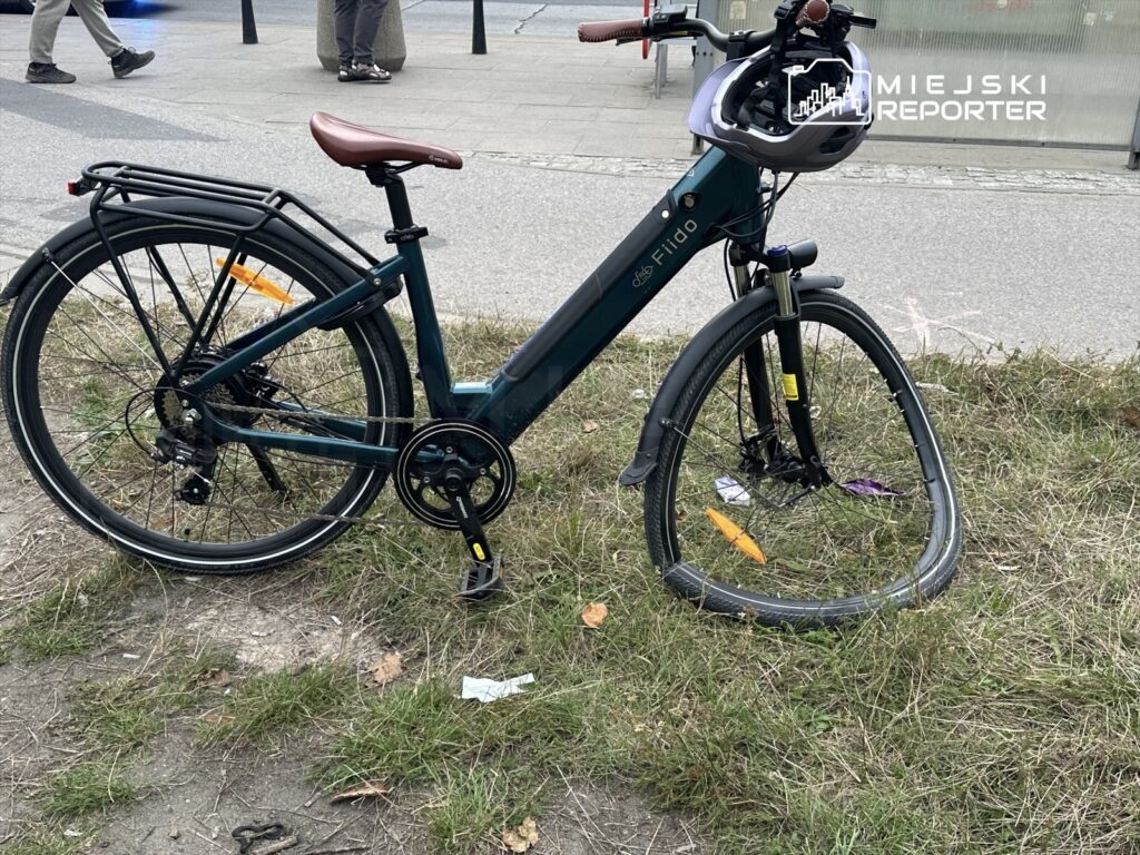 Zielony rower elektryczny stoi na trawie z uszkodzoną przednią oponą, w tle przechodnie na chodniku.