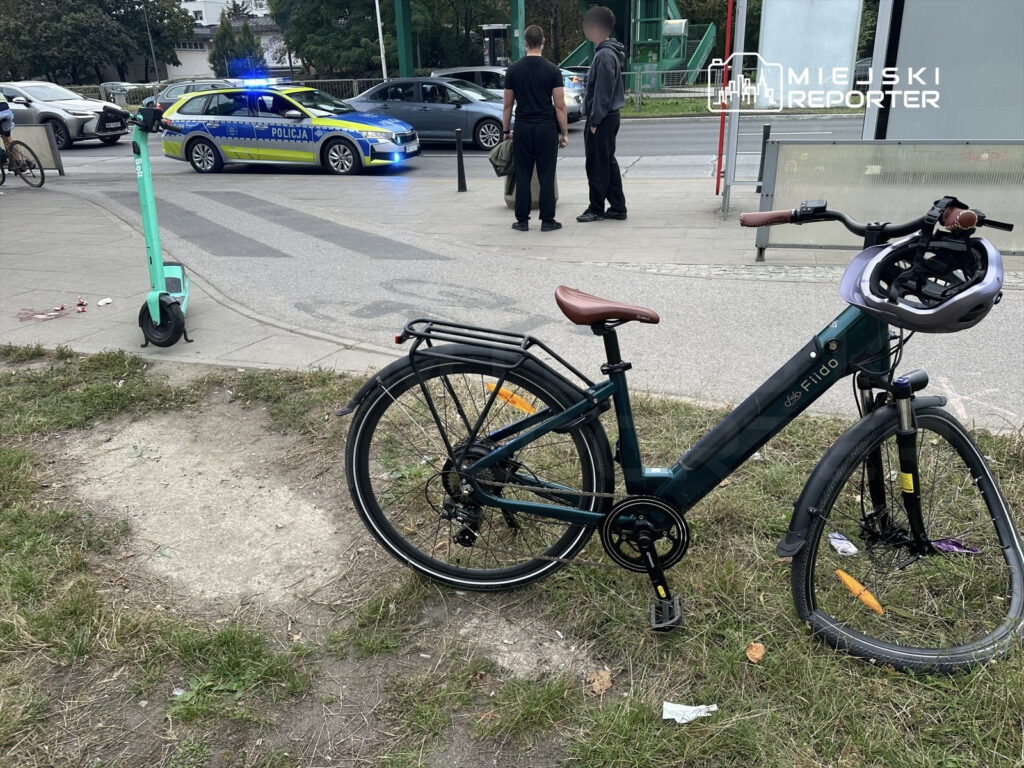 Policyjny radiowóz z niebieskimi światłami zatrzymany obok roweru na trawniku, w tle przechodnie i hulajnoga elektryczna.