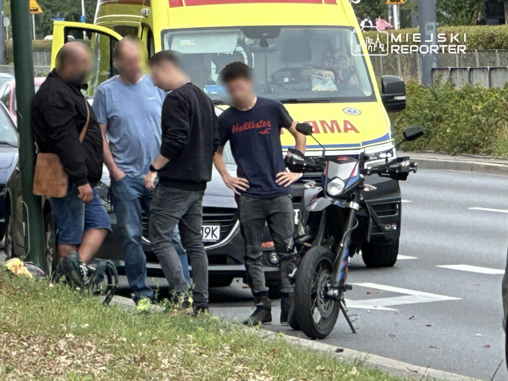 Czterech mężczyzn rozmawia obok motocykla przy drodze, w tle widoczna karetka pogotowia.