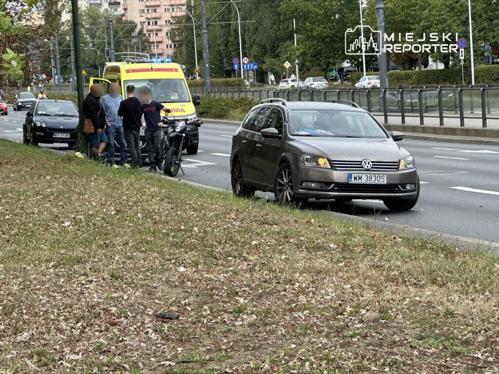Grupa mężczyzn rozmawia obok motocykla, podczas gdy w tle zatrzymuje się karetka na ruchliwej ulicy.