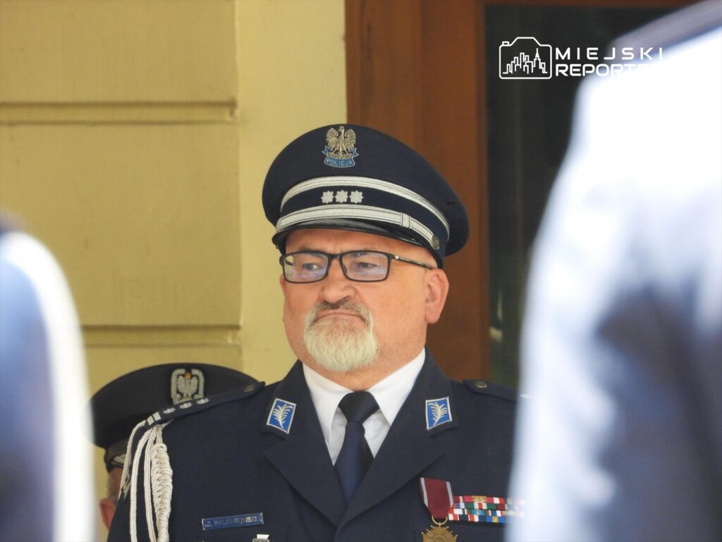Mężczyzna w mundurze policyjnym z odznaczeniami stoi w tle, obserwując ceremonię.