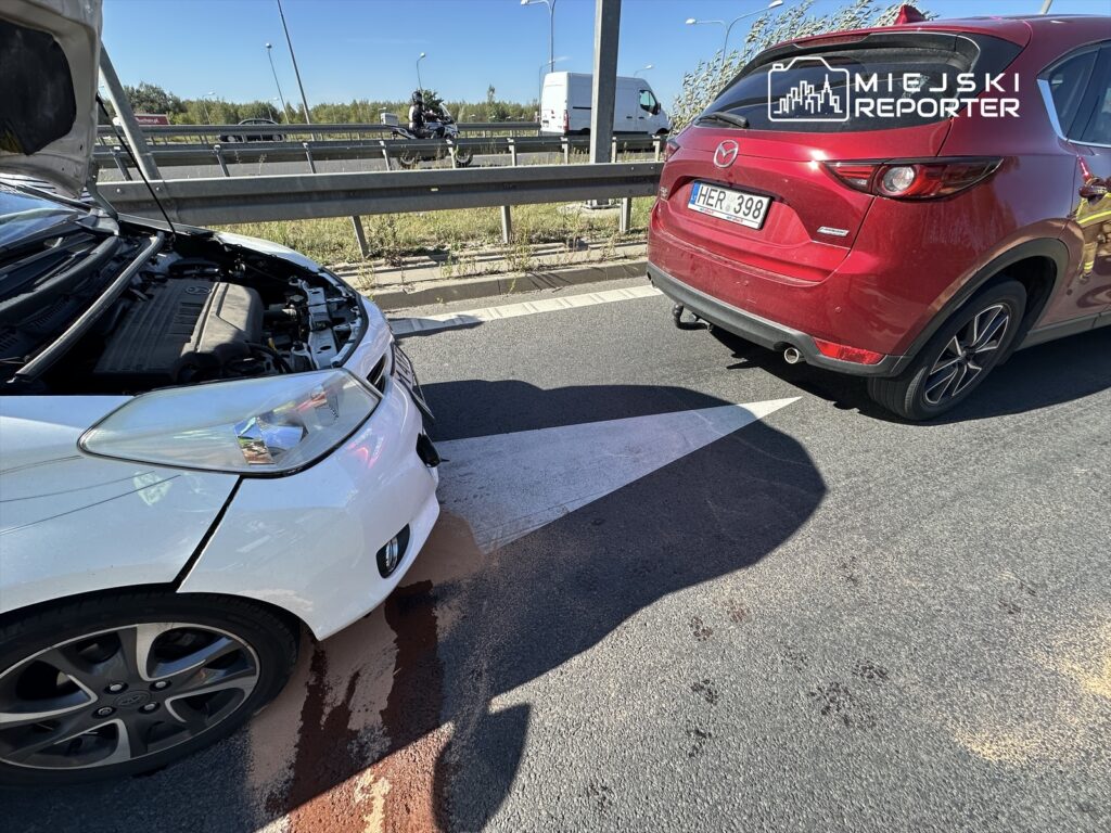 Biały samochód z otwartą maską stoi na pasie drogowym, obok niego czerwony SUV, a na jezdni widoczne plamy cieczy.