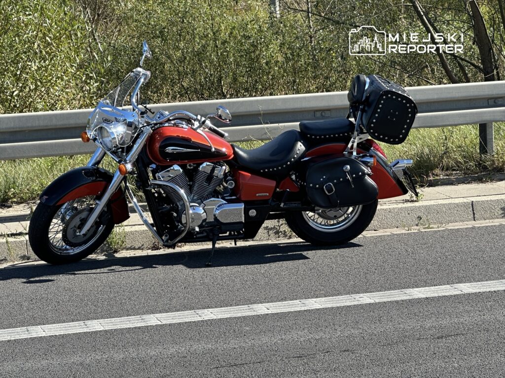 Motocykl w stylu cruiser o czerwonym kolorze zaparkowany na poboczu drogi, obok metalowej bariery.