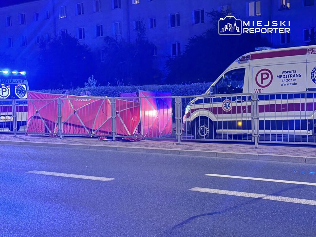 Na ulicy stoi ambulans z włączonymi światłami, obok ogrodzenie z czerwonymi osłonami, a w tle budynki mieszkalne.