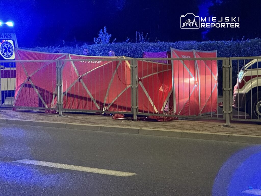Czerwone namioty medyczne ustawione przy ulicy, w tle widać ambulans z włączonymi światłami.