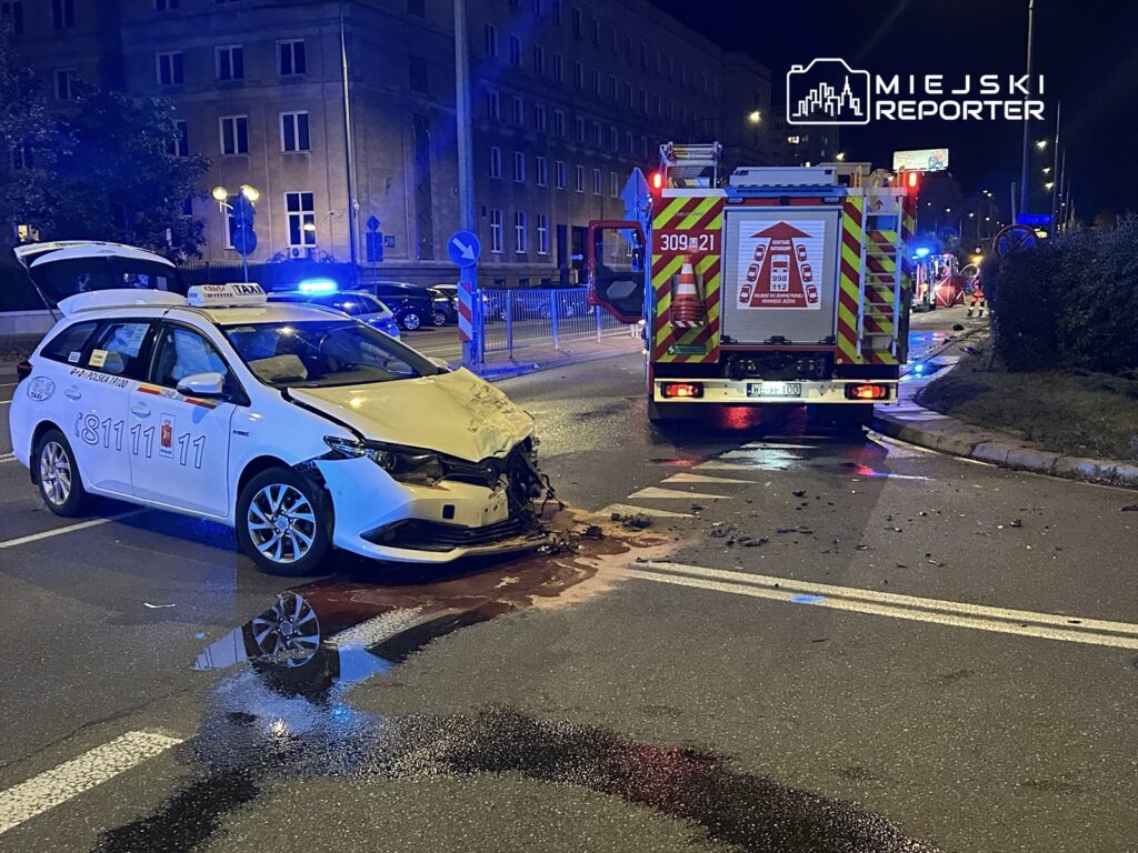 Uszkodzony samochód taxi z wgniecionym przodem stoi na jezdni, obok pojazdy straży pożarnej w nocy.