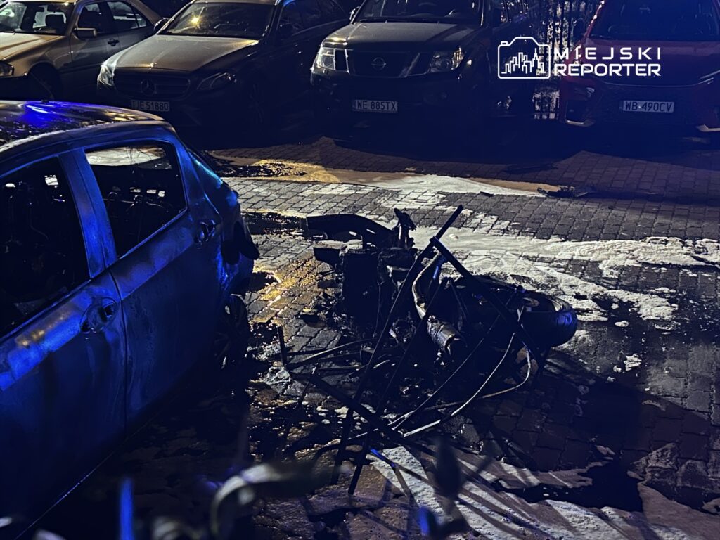 Spalony samochód osobowy stoi na ulicy, obok leży zniszczony motocykl, a w tle widać zaparkowane pojazdy.