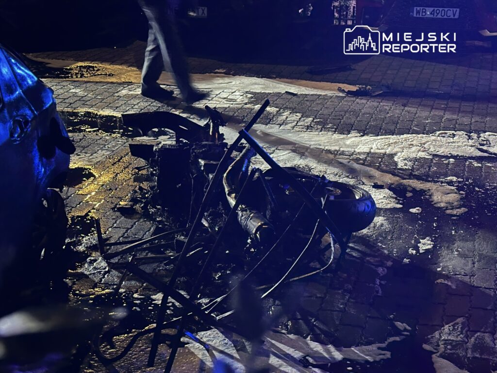 Zniszczony motocykl leży na ulicy, a obok przechodzi osoba w ciemnych spodniach. W tle widać odbicia niebieskiego światła.