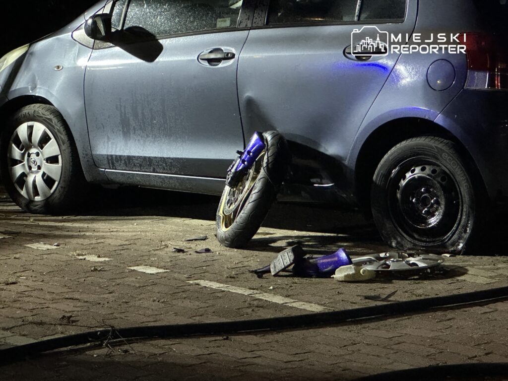 Uszkodzony samochód na parkingu z jednym zdjętym kołem i częściami leżącymi obok.