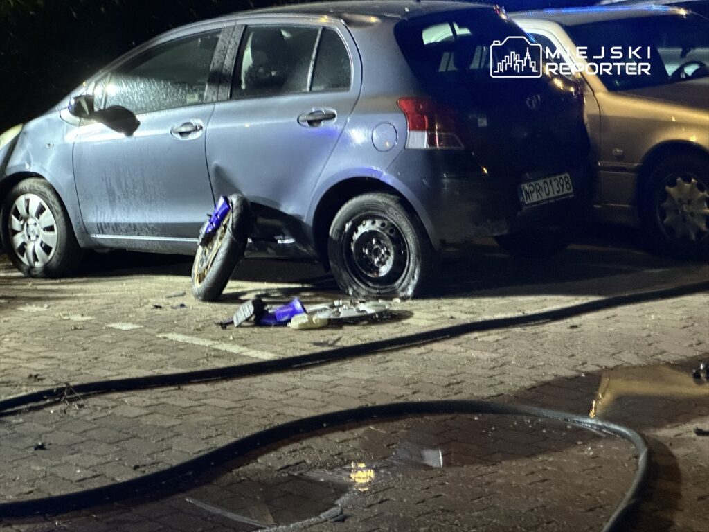 Uszkodzony samochód osobowy z odkręconym kołem na parkingu, obok leżące części motocykla.