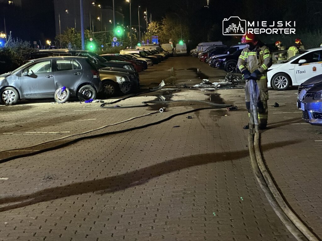 Strażacy w sprzęcie ochronnym rozkładają węże gaśnicze na parkingu, gdzie doszło do wypadku samochodowego.