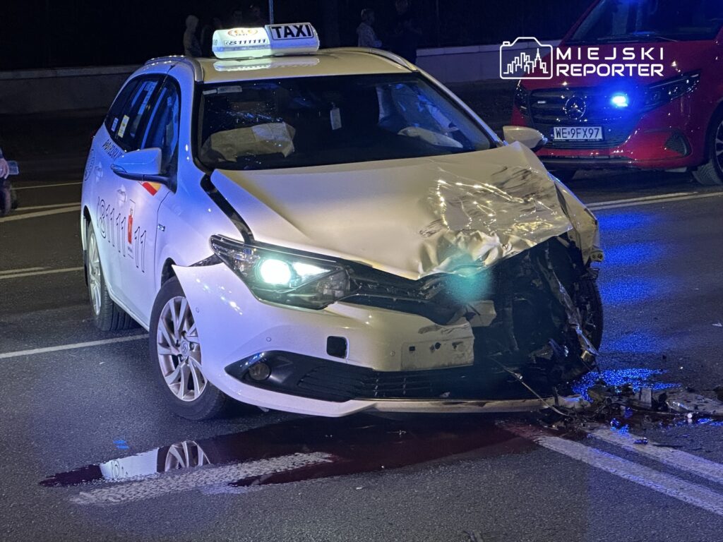 Rozbity samochód osobowy typu taxi z uszkodzoną przednią częścią na oświetlonej ulicy. W tle widoczny czerwony pojazd.