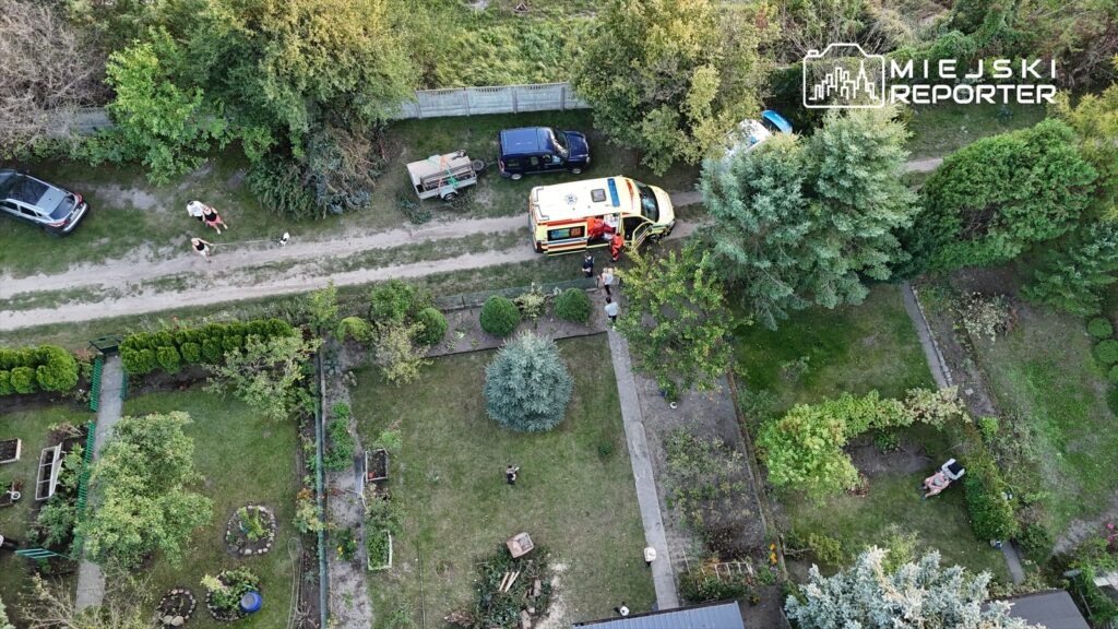 Karetka pogotowia zatrzymana na drodze, a obok niej grupa osób rozmawia w otoczeniu ogrodów.