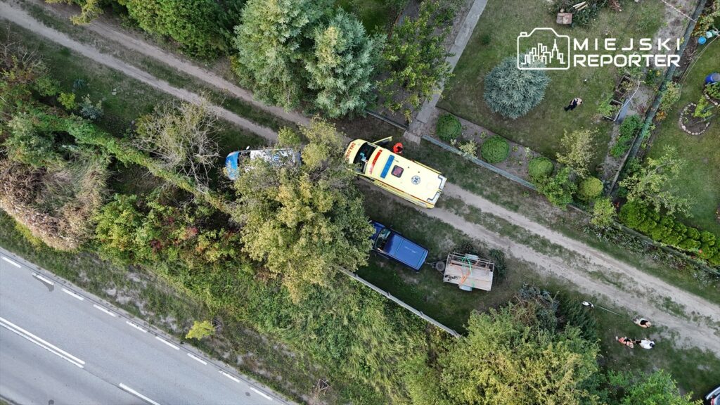 Karetka pogotowia parkuje na leśnej drodze między drzewami, obok dwóch samochodów osobowych. W tle widoczni mieszkańcy.