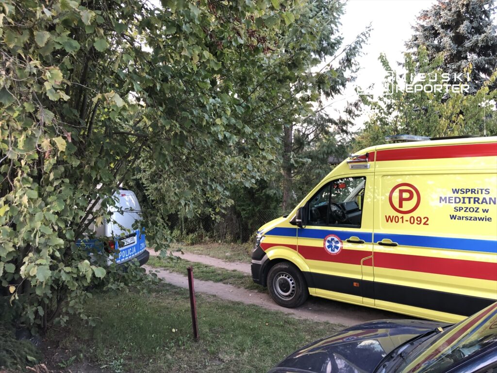 Karetka pogotowia medycznego zaparkowana na wąskiej drodze w otoczeniu drzew, obok nieoznakowany radiowóz policyjny.