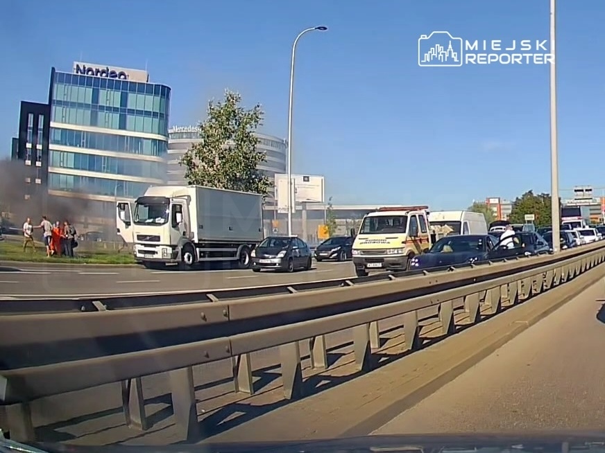 Na drodze widać samochody stojące w korku, obok grupę ludzi i pojazdy ratunkowe, a w tle dymi się z jednego z pojazdów.