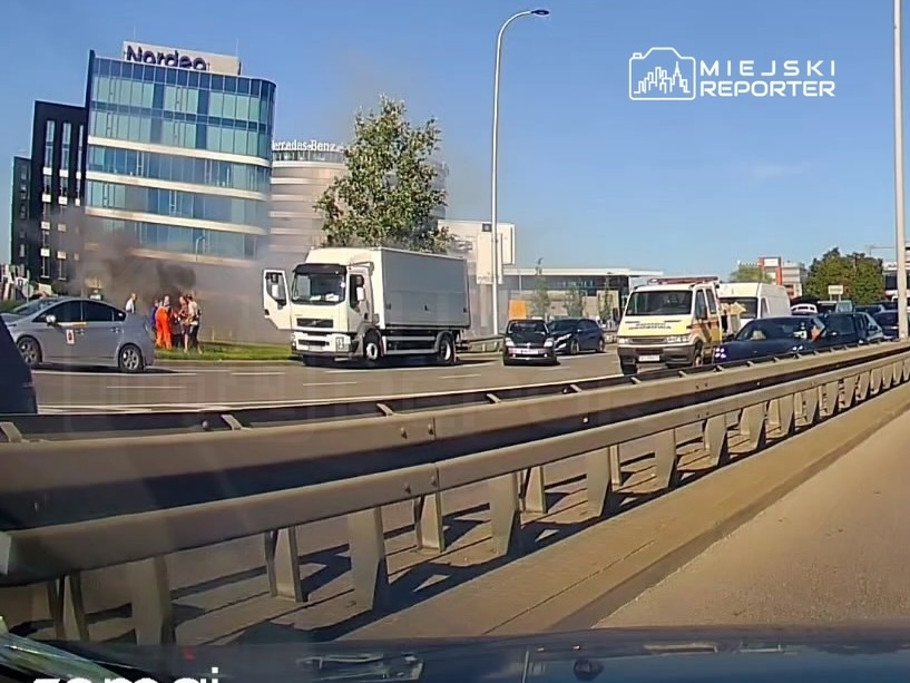 Grupa ludzi w pomarańczowych kamizelkach stoi obok płonącego pojazdu na poboczu drogi, w tle nowoczesny biurowiec.