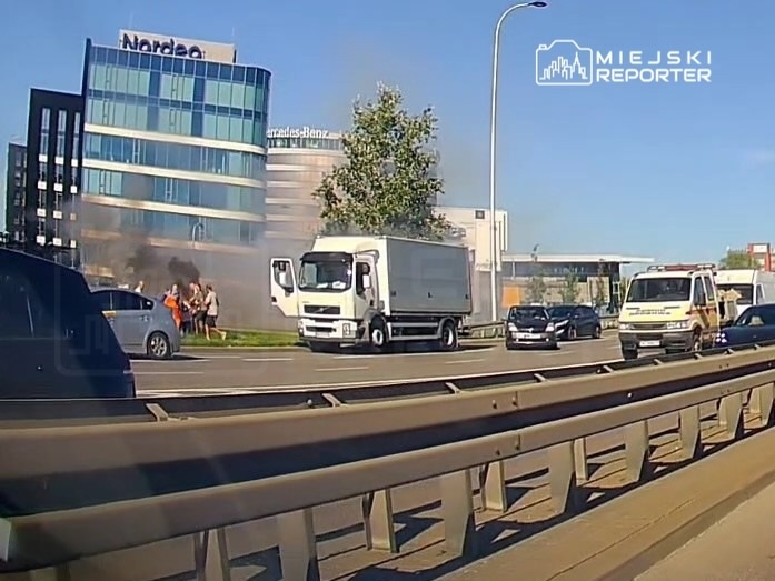Grupa ludzi obserwująca dym wydobywający się z samochodu ciężarowego na poboczu ruchliwej drogi, obok nowoczesnych biurowców.