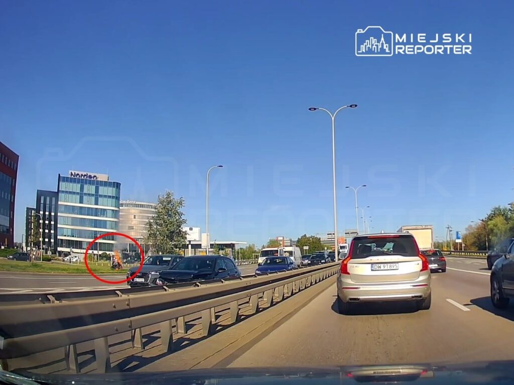 Samochody stoją w korku na drodze, obok nowoczesny budynek z przeszkloną fasadą i zielenią w tle.