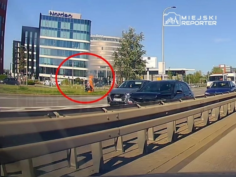 Motocykl objęty ogniem na trawniku obok nowoczesnego biurowca, w tle ruch uliczny.