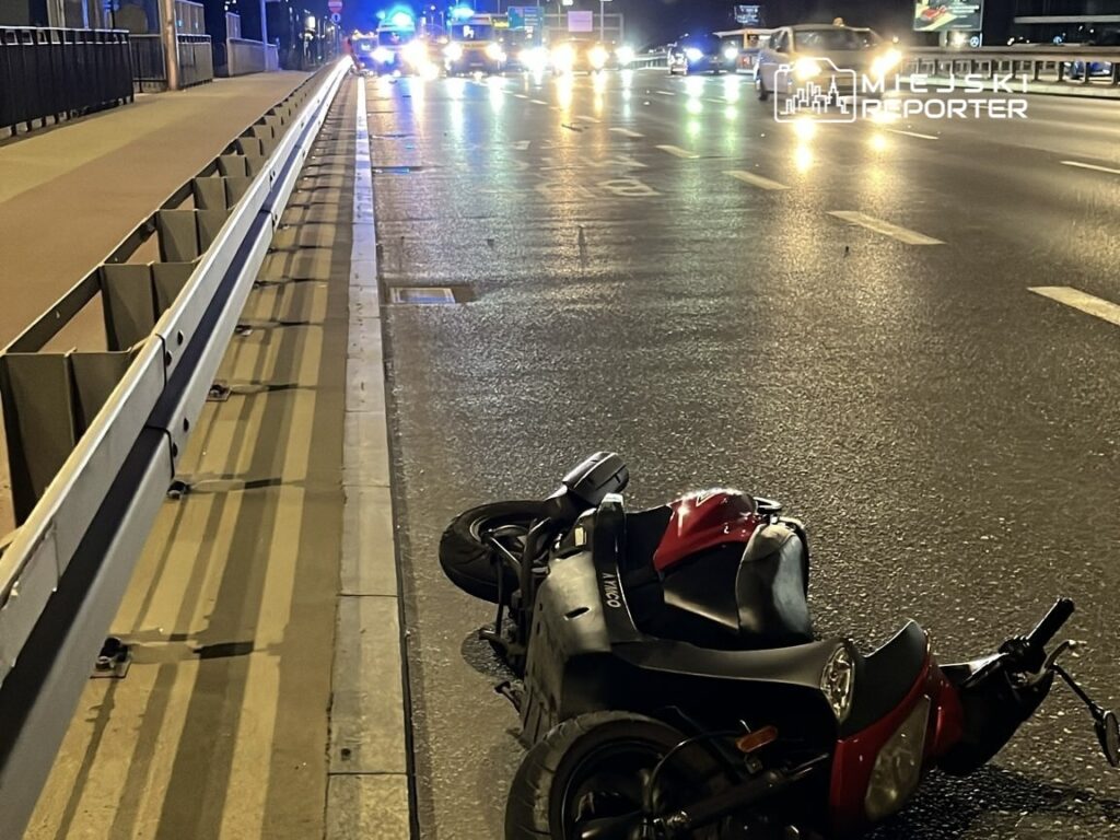 Motocykl leżący na boku na jezdni, obok widać pojazdy policyjne oraz światła w nocy.