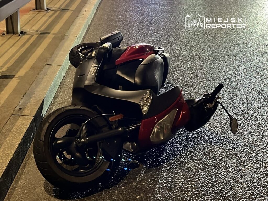 Przewrócony skuter leży na asfaltowej jezdni, obok krawężnika, w nocy.