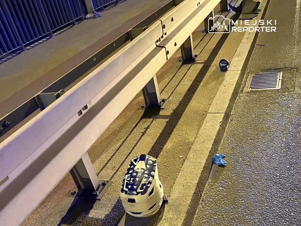 Na chodniku przy barierce leży biała torba oraz niebieska rękawiczka, w tle widoczna jest droga.