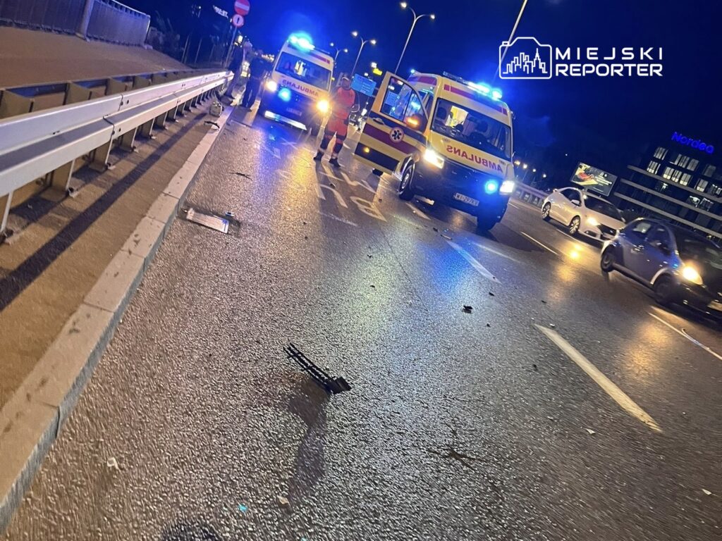 Na jezdni w nocy stoją dwa ambulansy z włączonymi sygnalizacjami świetlnymi, a obok pracują ratownicy.