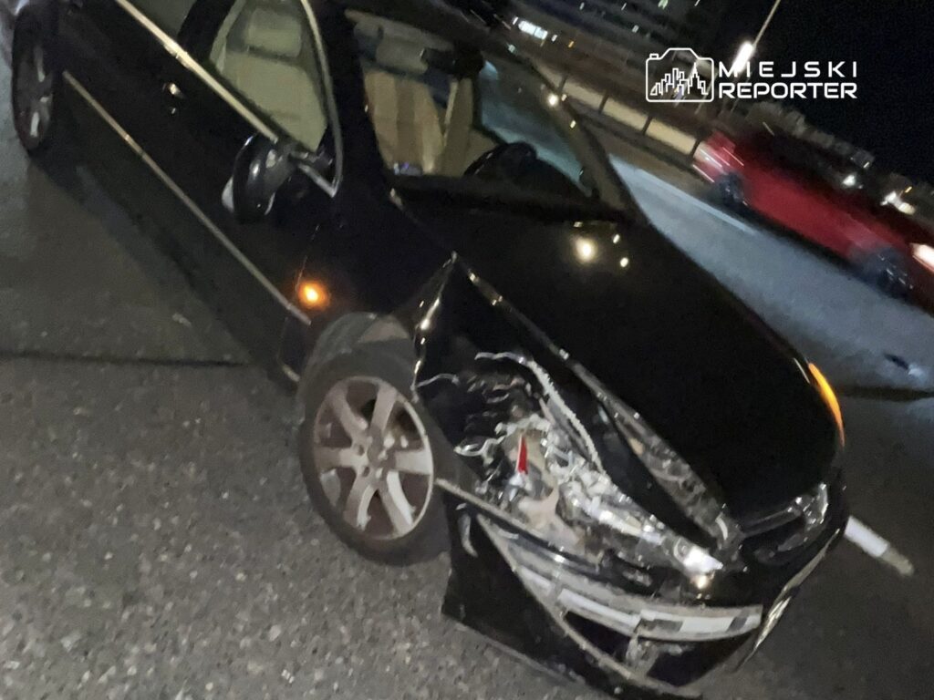 Czarny samochód osobowy z uszkodzoną przednią częścią stoi na drodze w nocy. W tle widać inne pojazdy.