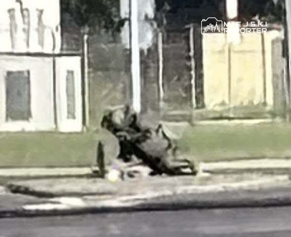Przewrócony skuter leży na chodniku obok drogi, w tle widoczne ogrodzenie i budynki.