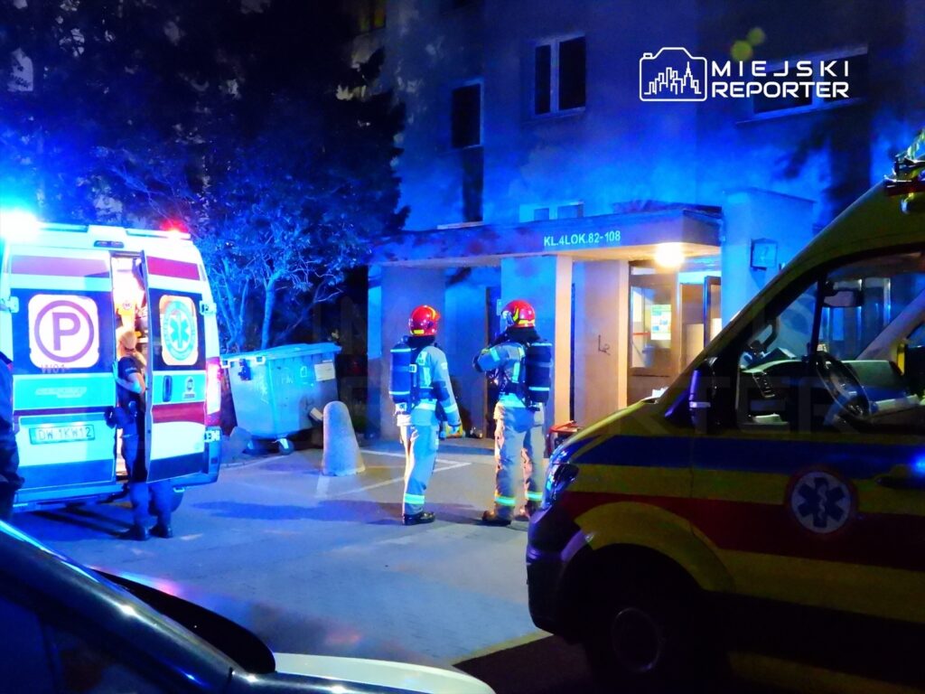 Ratownicy medyczni i strażacy w mundurach interweniują w nocy przed budynkiem mieszkalnym.