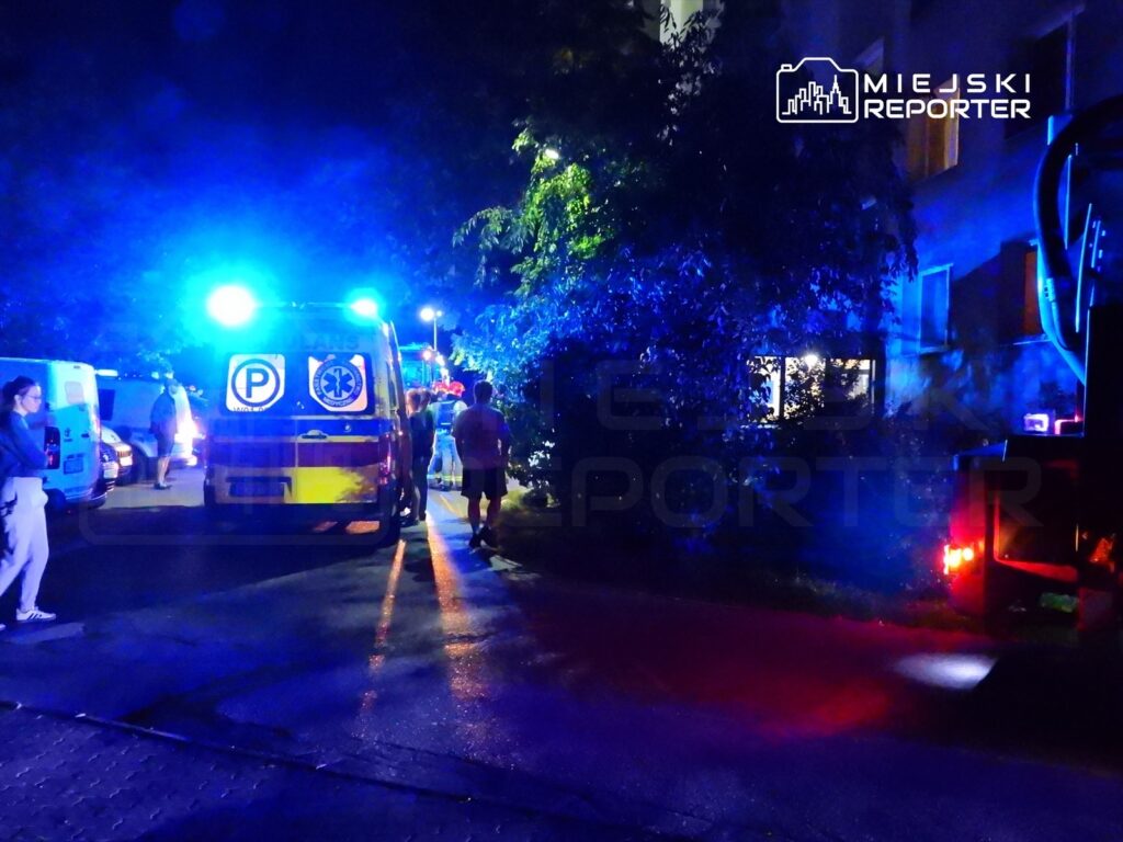 Karetka pogotowia z włączonymi sygnałami świetlnymi stoi na ulicy, obok grupy ludzi i pojazdów.