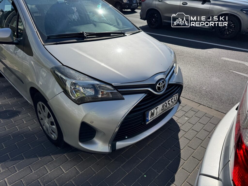 Srebrny samochód osobowy marki Toyota zaparkowany na chodniku w miejskim otoczeniu, obok innych pojazdów.