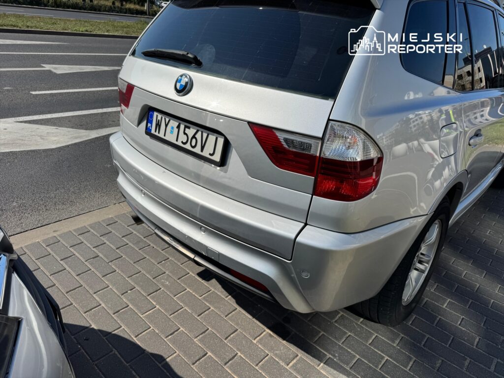 Srebrny samochód marki BMW zaparkowany na chodniku w miejskim otoczeniu, obok drogi.