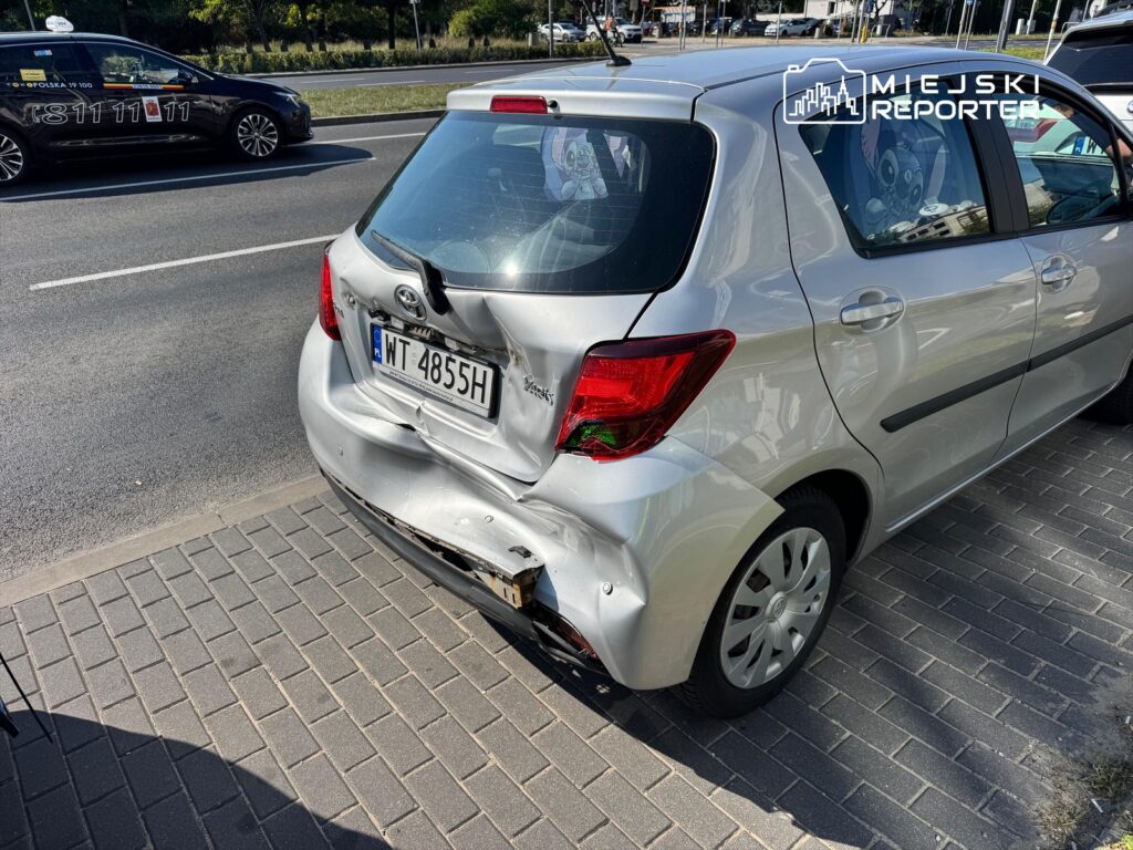 Uszkodzony tył srebrnego samochodu osobowego zaparkowanego na chodniku obok ruchliwej ulicy.